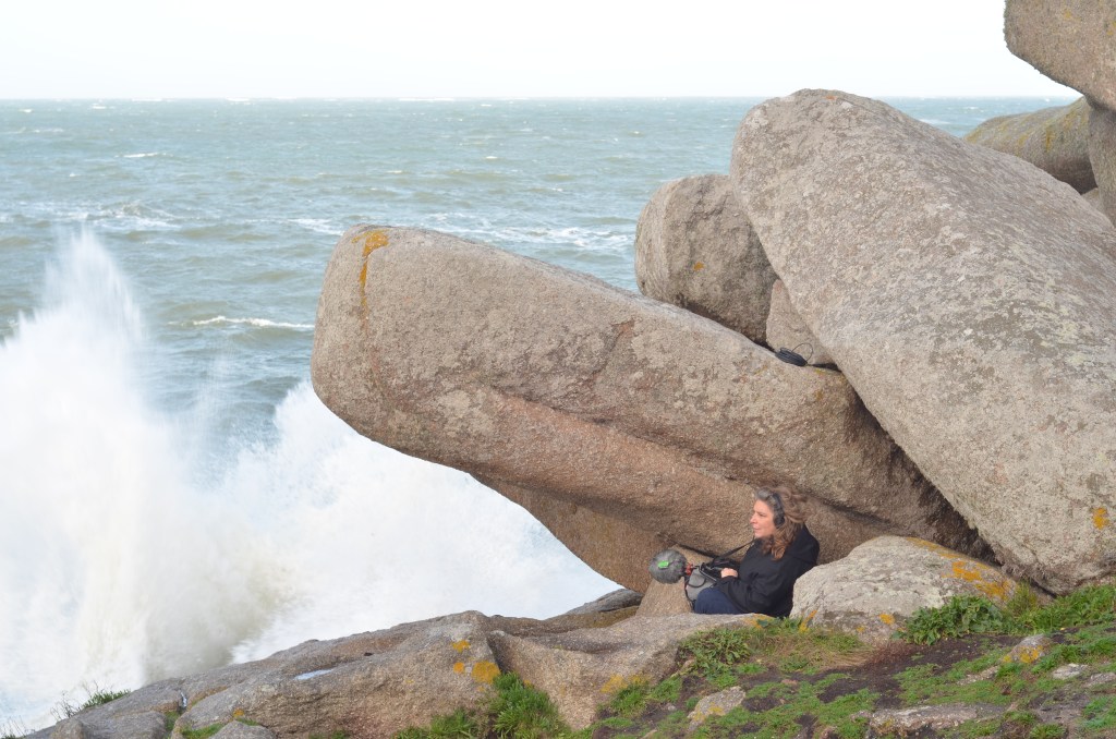A person sitting among large rocks by the sea, with waves crashing in the background.